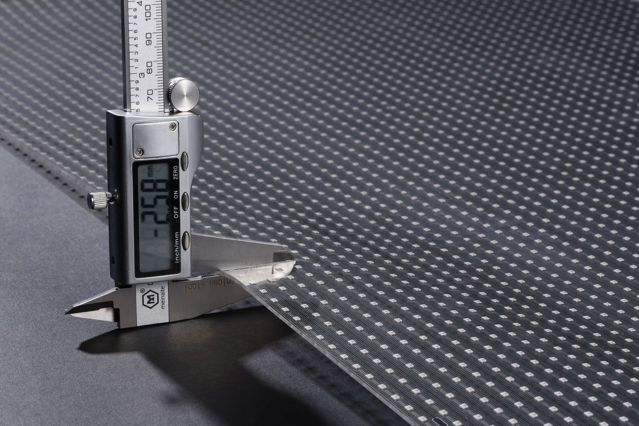 Digital calipers clamped on a sheet of transparent LED film reading 2.58 millimetres, with the LED grid visible across the panel
