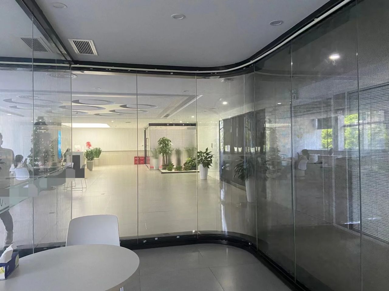 Curved glass office partition fitted with transparent LED, with the room, plants and people behind it clearly visible through the screen